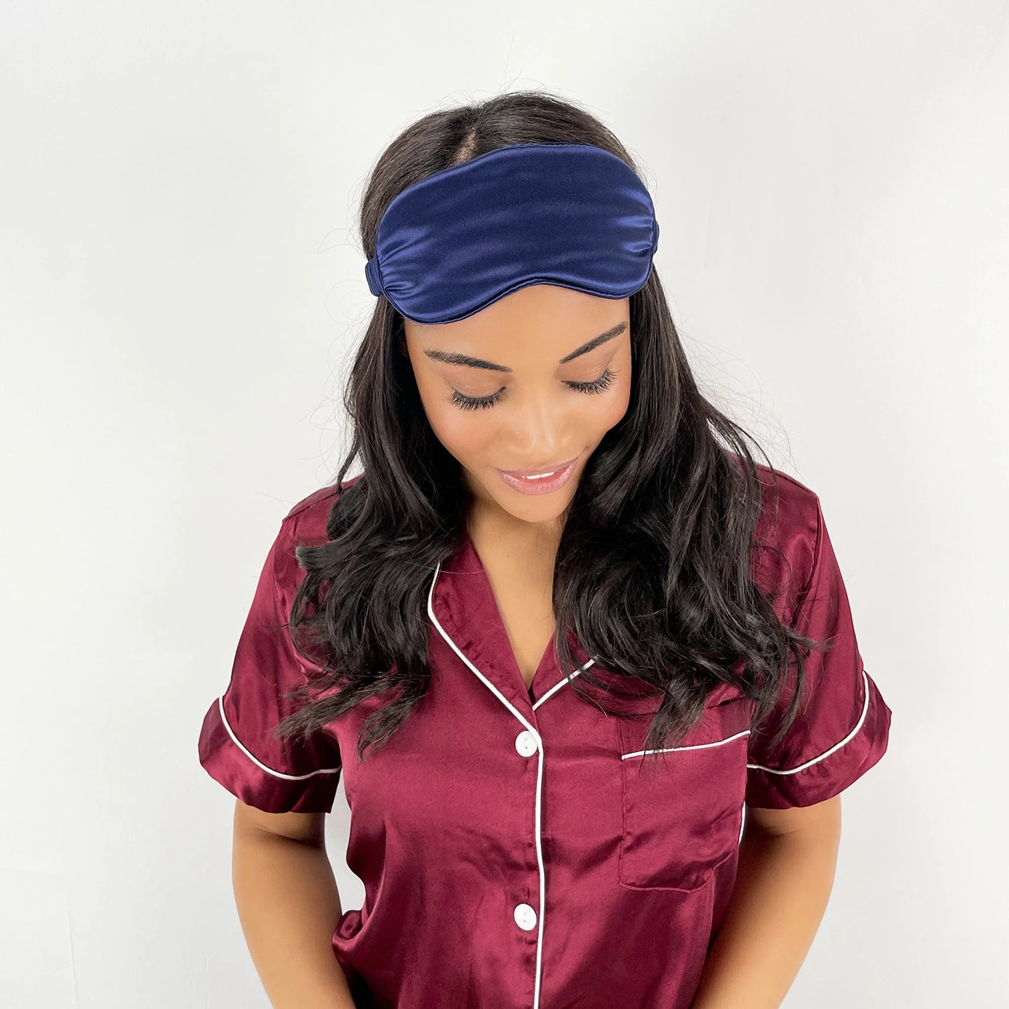 Woman wearing a maroon satin pajama set with white trim and a navy sleep mask.